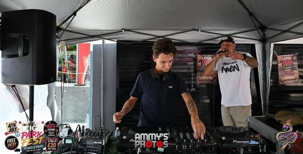 DJ at the mixing desk with a microphone performer behind him under a canopy.