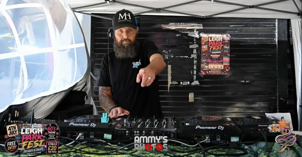 Bearded DJ pointing towards the camera from behind the mixing desk.
