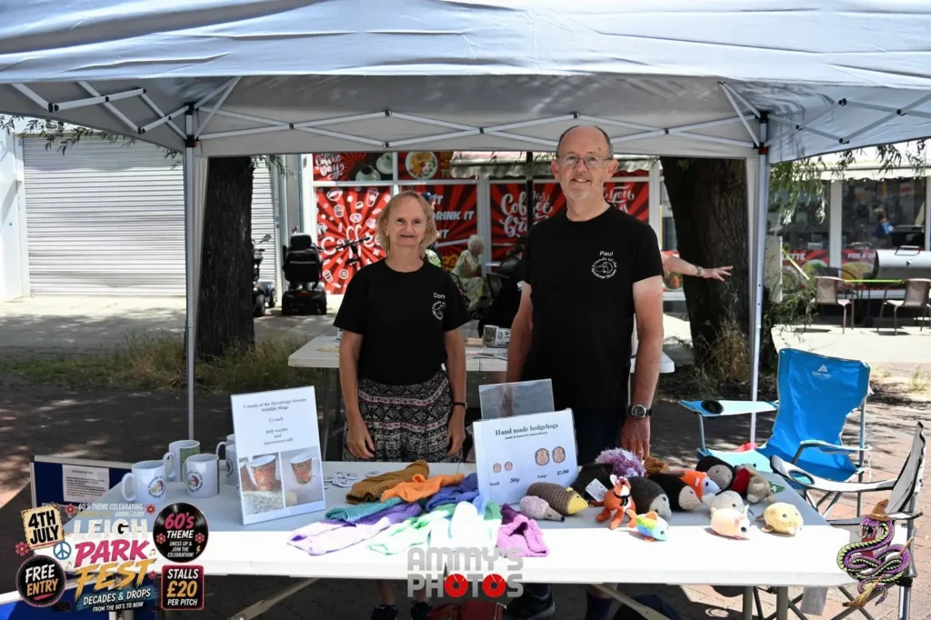 Two stallholders behind a table of colourful handmade items and mugs.
